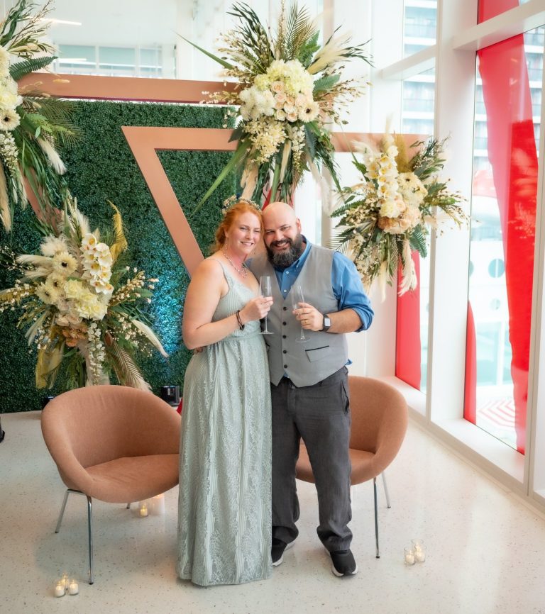 Bride & Groom celebrating their wedding ceremony officiated by Iris Ramil at Port of Miami Bride & groom celebrating their wedding, posing in front of floral decorations and greenery backdrop before going on Virgin Cruise.